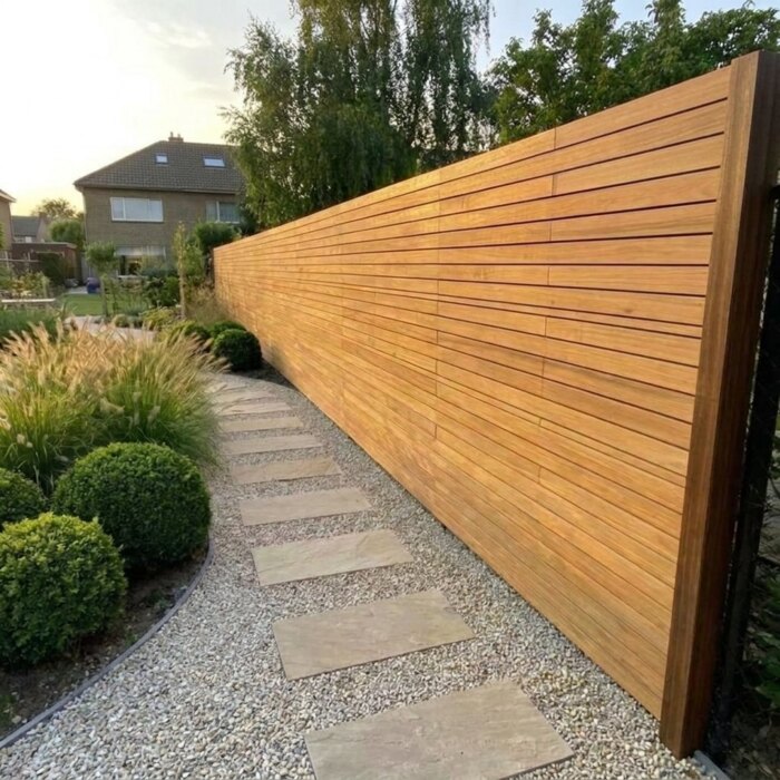 Lange teak schutting met 5 cm lamellen langs een tuinpad, strak in lijn geplaatst
