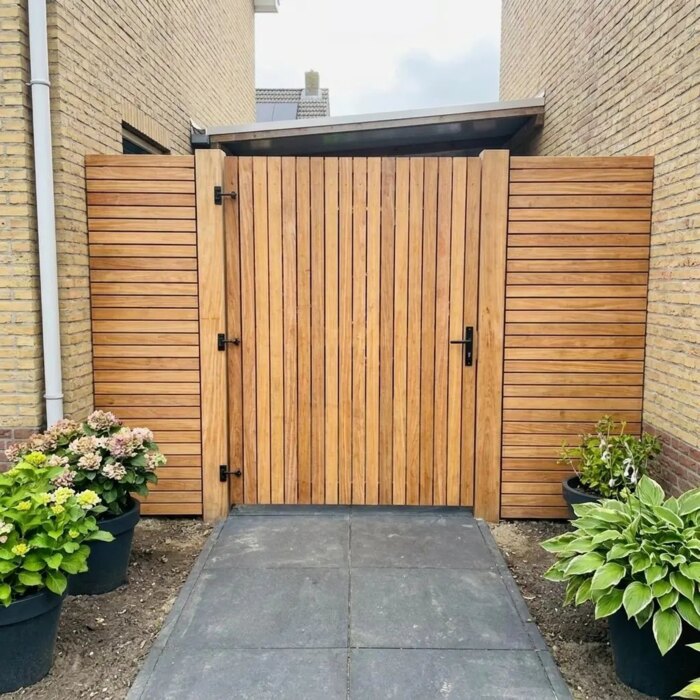 Teak tuinpoort met lamellen van 5 cm in een zijpad naast een huis van gele baksteen
