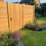 Teak tuinpoort van 5 cm lamellen in een teak schutting achter een border met siergrassen