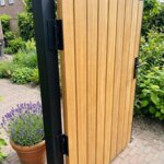 Detail van het zwarte scharnier aan de teak tuinpoort met lamellen van 7 cm