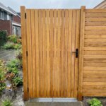 Teak tuinpoort met brede lamellen van 9 cm na een regenbui, naast een teak schutting